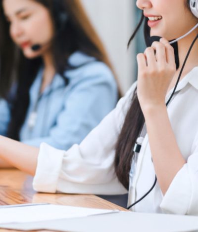 Two BPO customer service agents wearing headsets at a desk, engaged in conversation, with office supplies visible
